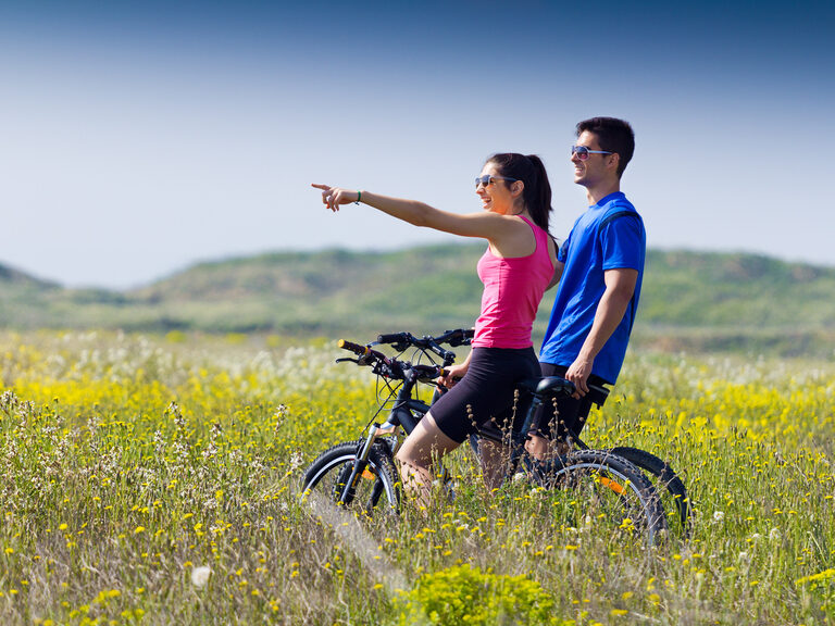 Junges, sportliches Pärchen auf Fahrrädern bei einem Halt auf einer grünen Wiese. Junge Frau schaut in den Wald während Sie auf dem Fahrrad Pause macht.