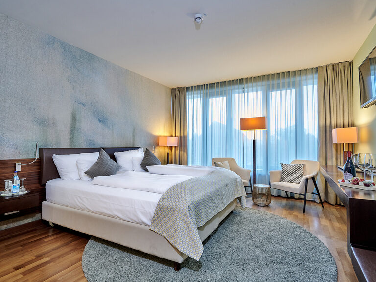 Ein Hotelzimmer mit einem breiten Bett und einem an der Wand montierten Fernseher. Darstellung eines geräumigen Schlafzimmers vor einer Balkonfront.