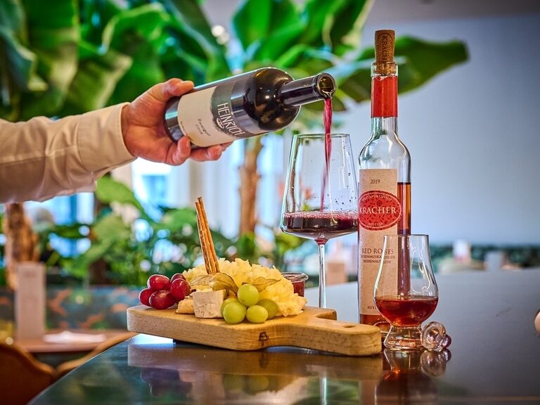 Ein Käsebrett und Wein, der gerade in ein Glas geschenkt wird an der Bar im Hotel ASAM. Junge Frau schaut in den Wald während Sie auf dem Fahrrad Pause macht.